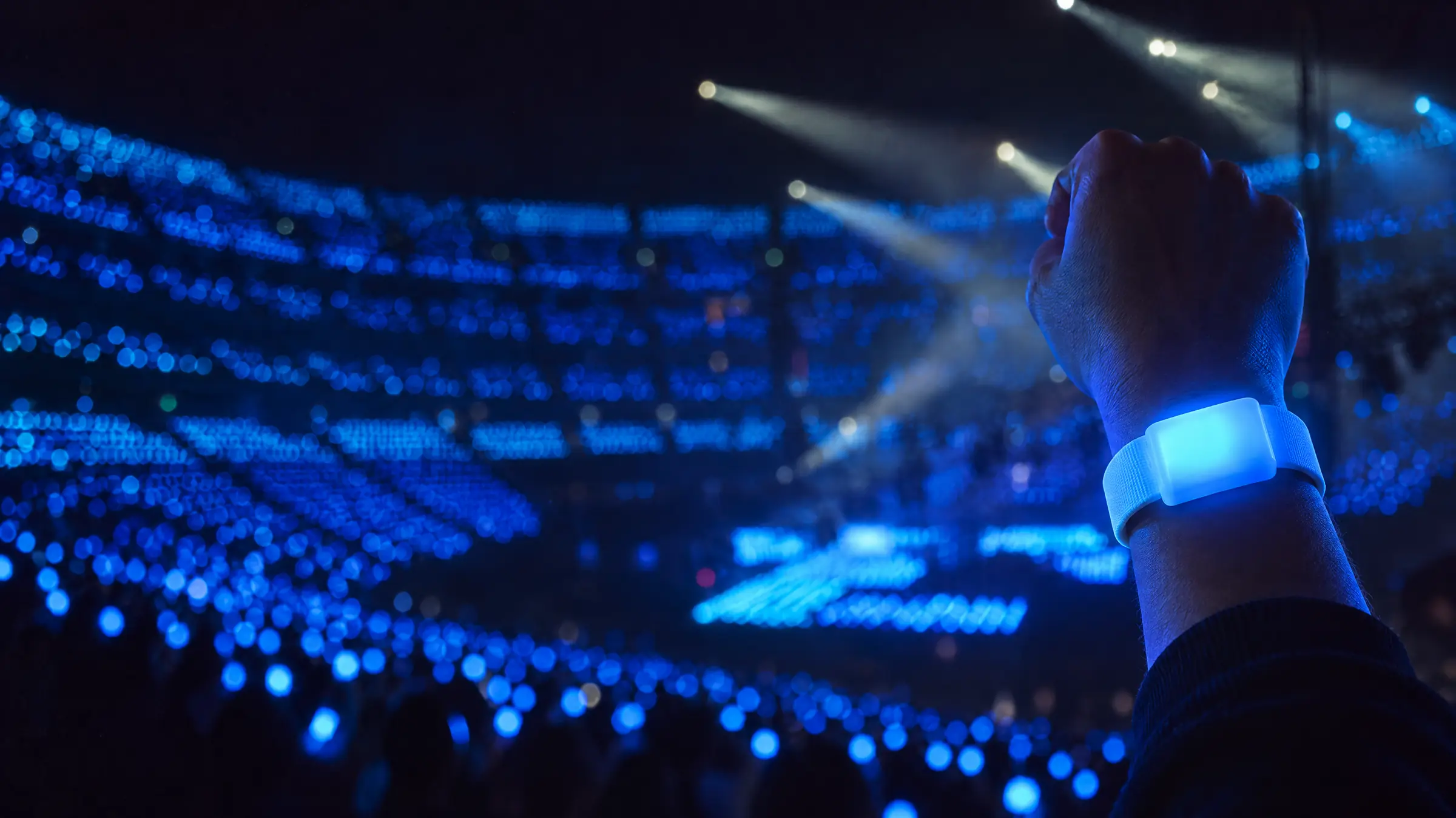 A raised arm wearing a rechargeable LED wristband glowing blue, in front of a stadium where every wrist glows the same blue
