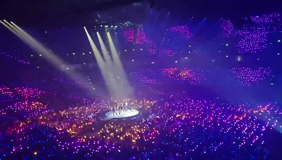 An arena concert seen from the top tier, the audience lit in purple and amber wristbands around a round stage