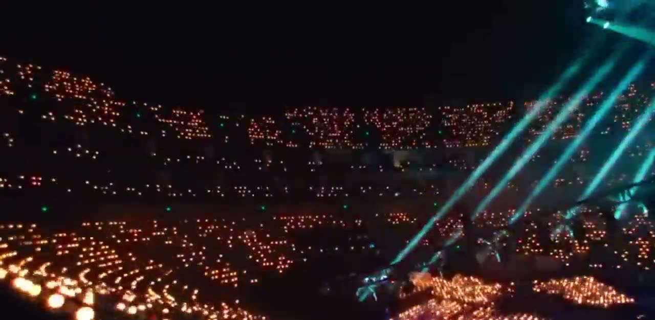 A full stadium at night, thousands of wristbands glowing in the stands while beams sweep the stage